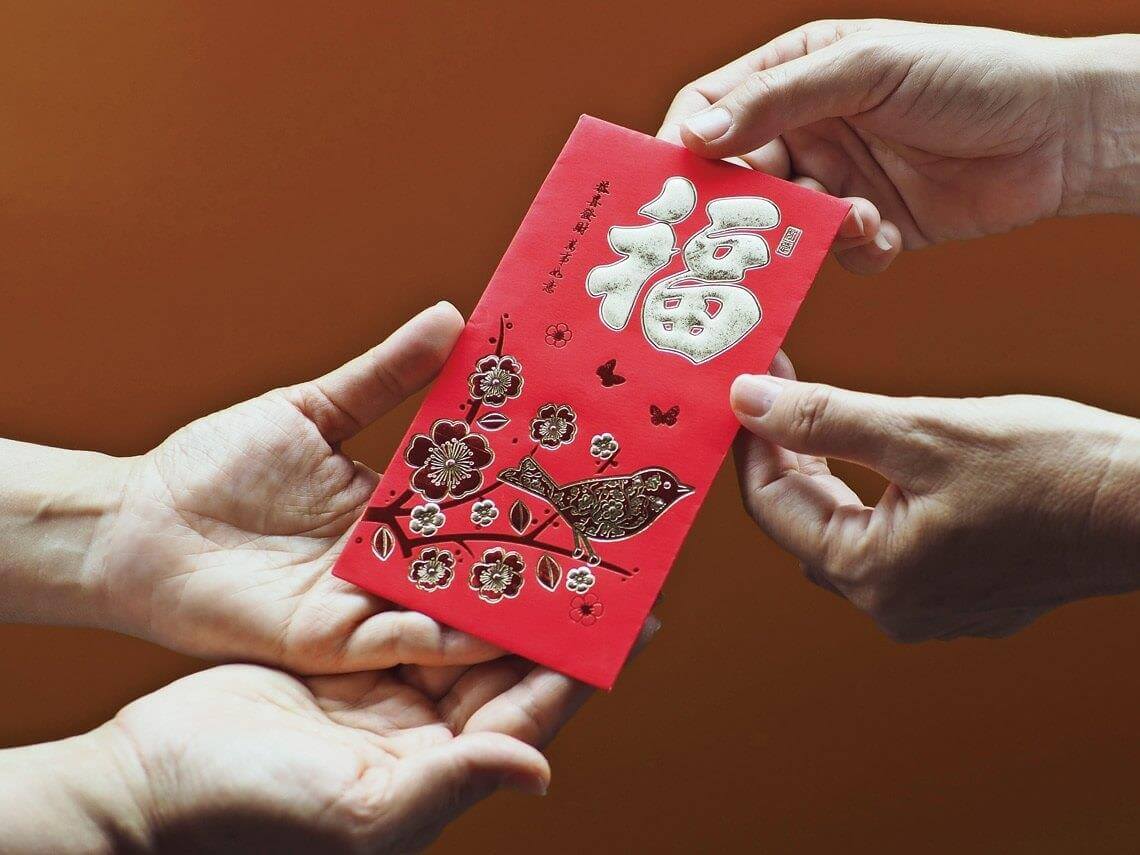 Giving Red Envelopes on the Occasion of the New Year 12 Three hands exchange a red envelope, intricately designed with florals and birds, along with Chinese characters, against a warm orange background—a seamless blend of tradition and meticulous craftsmanship akin to civil engineering outsourcing.